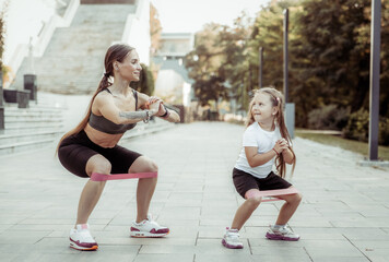 An athletic mother trains with her daughter. Workout with fitness rubber bands outdoors, healthy...