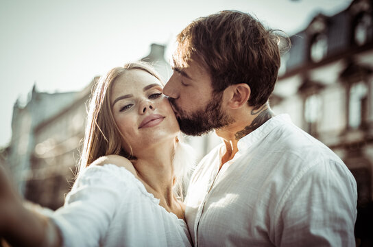 Selfie portrait of a young couple in love. Man kisses his girlfriend on cheek in city