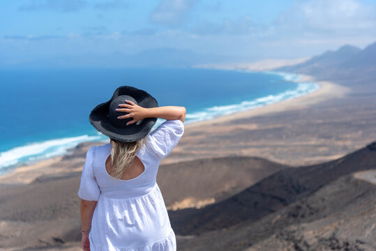 Joven rubia de pelo largo vestida de blanco sosteniendo un sombrero de paja de espaldas disfrutando de la playa de arena blanca y aguas cristalinas de la playa de Cofete en Fuerteventura, Canarias