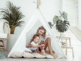 Mom and daughter are playing with a developing pyramid while sitting in wigwam in modern minimalistic light room with house plants © splitov27