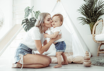 Maternal love concept. Mom hugs and kisses her little daughter on the cheek in room with wigwam © splitov27