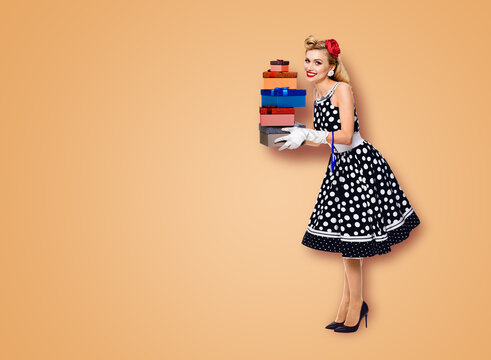 Big sales discounts, rebates offers ad concept - full body image of happy smiling beautiful woman in pin up black dress with polka dot, white gloves, hold gift boxes, isolated latte beige background