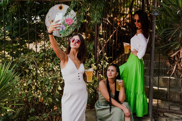 Three stylish young women dancing and laughing with drinks in hand against a colorful graffiti wall. A lively urban summer vibe full of joy, friendship, street art, and carefree energy.