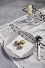 Table Easter decor set with quail eggs on plate, willow branches and candles on white embroidered table cloth.
