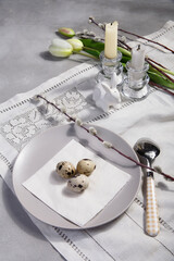 Table Easter decor set with quail eggs on plate, willow branches, white tulip and candles on white embroidered table cloth.