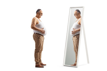 Full length profile shot of a mature man in an underwear vest looking at his big belly in a mirror - obrazy, fototapety, plakaty