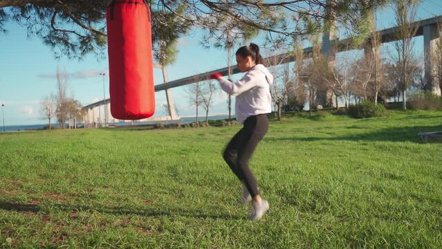 Female boxer dreesd hoodie warming up body muscule. Athletic female prepares for boxing workout outdor in park with punching bag. Fighter warm up body before training Female fitness motivation. 