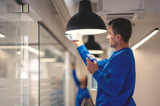 Professional hygiene maintenance. A cleaner cleans the chandelier in the office while in the background another cleaner washes the glass panels