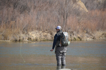 A single man fly fishing on a river with a hat and backpack.