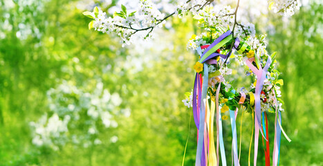 Spring floral wreath with colorful ribbons on tree in garden, green natural background. floral decor. Symbol of Beltane, Wiccan Celtic Holiday beginning of summer. witchcraft ritual. banner