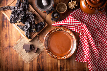 Empty Mexican clay plate, set with some typical ingredients of Mexican cuisine, pasilla chili, chocolate, molcajetes and clay pot on rustic wooden table, top view.
