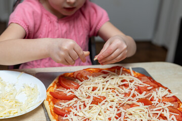 Little girl making pizza with cheese. Small child cooking meal, sitting at kitchen table at home.