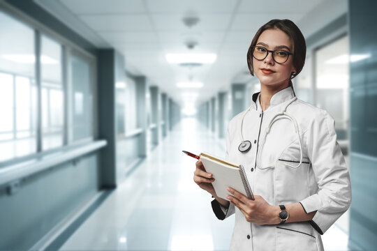 Portrait of surgeon woman dressed in white robe posing against corridor of hospital.