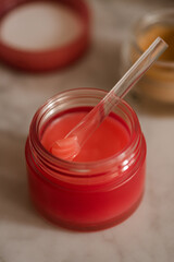 Red plastic jar and spoon with lip balm product close up. Health and skin care morning routine. Selective focus.