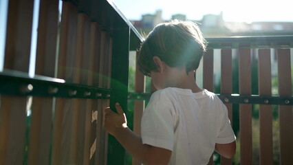 One small boy standing by balcony fence staring outside from second floor with lens flare sunlight. Child holding in wooden bars looking out