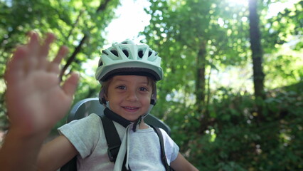 One happy child waving hello seated in bike seat wearing helmet protection. Adorable kid waves with hand in bicycle chair