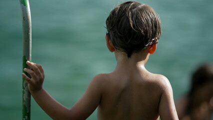 One contemplative small boy. Back of wet shirtless child standing outside holding into metal bar by water. Thoughtful little kid stands outdoors
