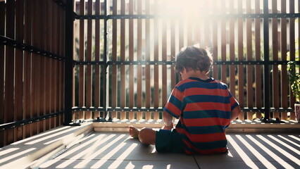 One child sitting at balcony in morning sunlight with flare. Small boy in apartment terrace