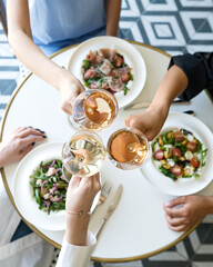 Three women are having lunch in restaurant and drinking wine. Glasses of white wine in hands. Three plates of salad on round white table. Light lunch. Italian restaurant. Healthy food. Top view. 