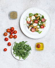 Plate of salad with arugula, tomatoes, mozzarella and chicken. Salad, olive oil, spices, tomatoes and arugula on white table. Mediterranean cuisine. White background. Top view. Copy space.
