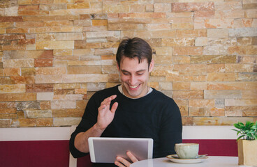 Young and handsome man talking in a videoconference media in a tablet in a coffee shop while having a coffee. High quality photo
