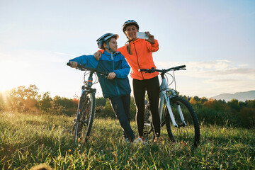 Mother and son ride a bike. Happy cute boy in helmet learn to riding a bike in park on green meadow in autumn day at sunset time. Family weekend.