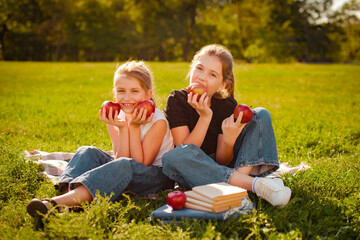 two girls taste delicious apples on green grass in park or school yard doing homework after classes