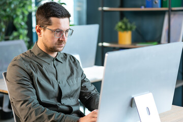 Serious handsome bearded man in casual shirt sitting on workplace and working with computer