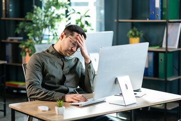 Young exhausted business man suffering from headache in modern office with computer on desk.