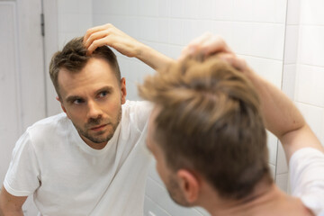 Middle aged caucasian white man with a short beard looks at his hair in the mirror in the bathroom and worried about balding. The concept of the problem of male hair loss, early baldness and alopecia - obrazy, fototapety, plakaty