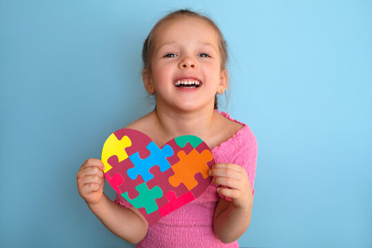 A little laughing girl in a pink T-shirt shows a postcard with a picture of puzzles. A symbol of awareness about autism. The social problem of communicating with special people