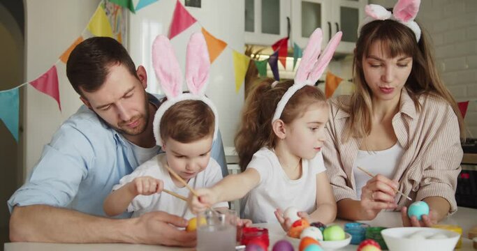 Joyful family of four wearing bunny ears painting Easter eggs together at the table in bright modern kitchen. Family gathering in preparation for Easter.
