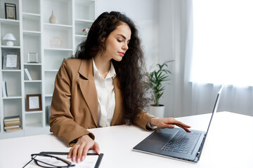 A young curly-haired brunette girl works at a laptop. Freelancer working from home, remote work. Successful manager girl, in a white office.