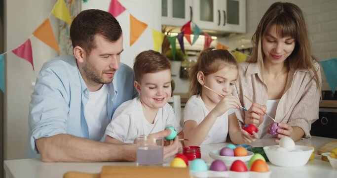Joyful family of four painting Easter eggs together at the table in bright modern kitchen. Family gathering in preparation for Easter.