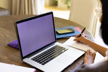 Focused biracial woman sitting at table and using laptop with copy space in living room