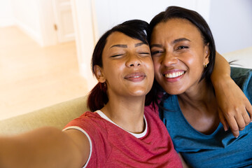Portrait of happy biracial sisters looking at camera, taking selfie, smiling and embracing - obrazy, fototapety, plakaty