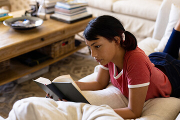 Happy biracial woman lying on couch and reading book