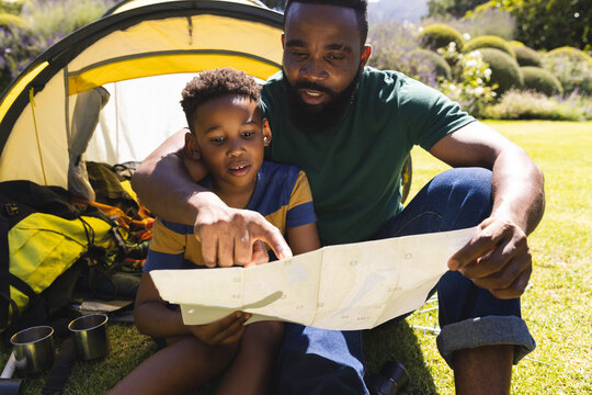 Happy african american father and son sitting on grass and reading map next to tent in garden