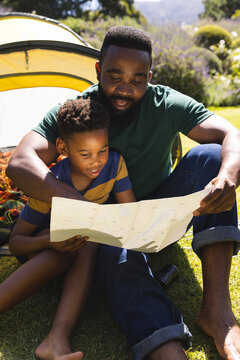 Happy african american father and son sitting on grass and reading map next to tent in garden