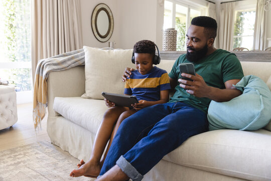Happy african american father and son sitting on sofa, using tablet and smartphone