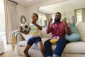 Happy african american father and son sitting on sofa, watching football match and supporting