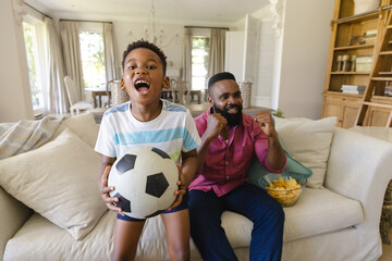 Happy african american father and son sitting on sofa, watching football match and supporting