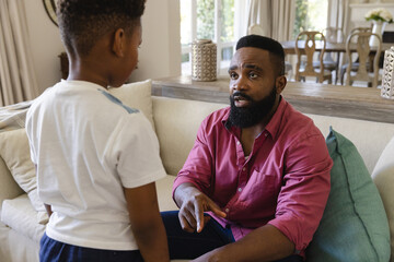 African american father sitting on sofa in living room, disciplining his son