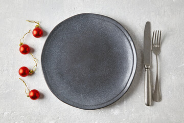 An empty gray ceramic plate with cutlery surrounded by red Christmas balls on a light concrete...
