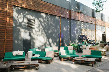 Outdoor patio. Furniture from pallets, with green pillows, luminous garlands against a concrete wall. Wedding party on the street.