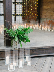 The wooden patio is adorned with white candles in glass sconces, a bouquet of green leaves and twigs in an antique copper vase. Garlands glow in the background.