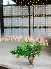 The wooden patio is adorned with white candles in glass sconces, a bouquet of green leaves and twigs in an antique copper vase. Garlands glow in the background.