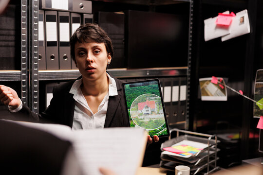 Woman Investigator Showing Crime Scene Photos On Tablet Computer, Discussing Criminal Investigations With Police Officer. Private Detectives Working Late At Night In Dark Room