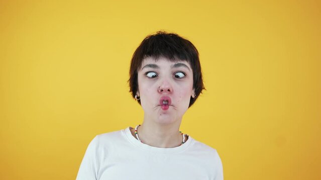 Surprised funny young woman poses indoor against yellow studio background looking at camera with playful fish face grimace pout lips. Copy space. High quality 4k footage