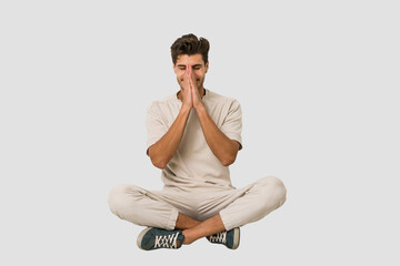 Young caucasian man sitting on the floor isolated on white background holding hands in pray near mouth, feels confident.